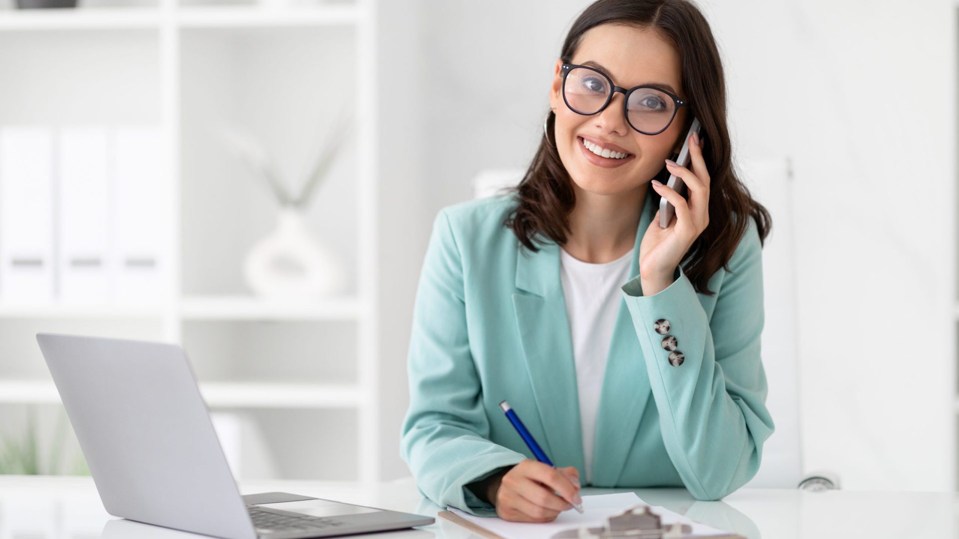 Smiling millennial caucasian lady secretary in suit and glasses calls by phone at workplace in light office interior. Business with device, communication with client, work, multitasking manager